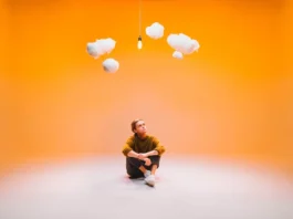 Devin Kennedy In My Imagination shot by Caden Huston orange gradient background with artist Devin Kennedy sitting and looking up at some illustrated 3D clouds